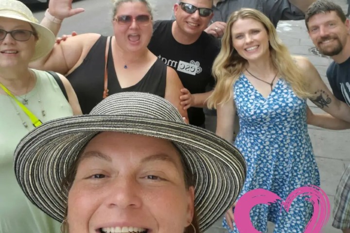 Group of people smiling and posing on a street with a pink heart graphic.