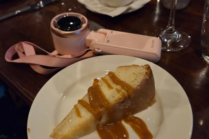 Cheesecake slice with caramel sauce on a plate, beside a camera and glass.