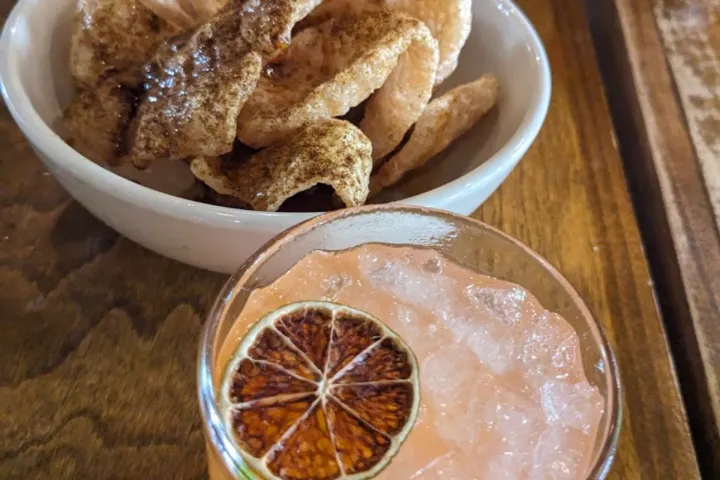 Glass with cocktail and lime slice, bowl of chips on a wooden table.