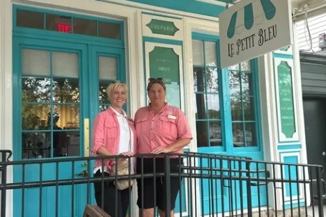 Two people stand in front of Le Petit Bleu cafe with turquoise doors and a hanging sign.