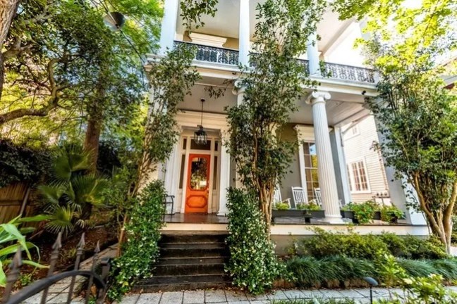 Front view of a two-story house with white columns and a wooden door, surrounded by greenery.