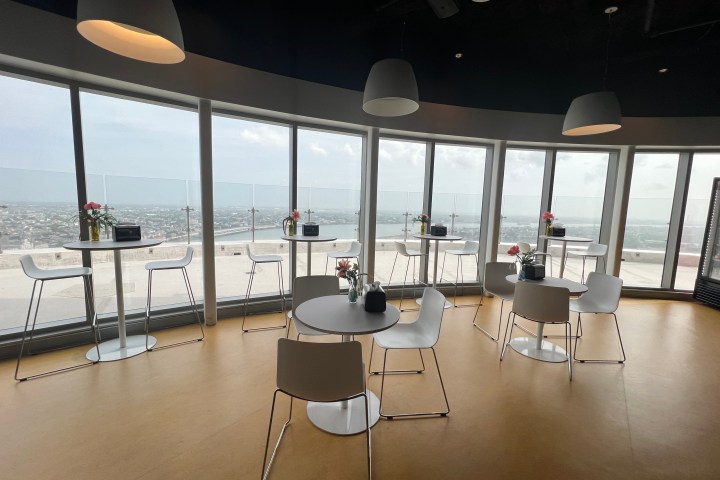 Modern café interior with tall windows showing city view, round tables, white chairs, and hanging lamps.