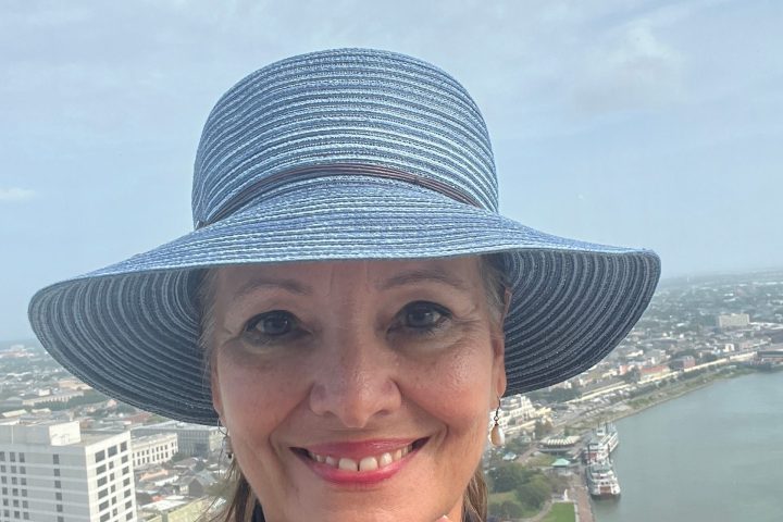 Woman in blue hat takes a selfie with a cityscape and water in the background.