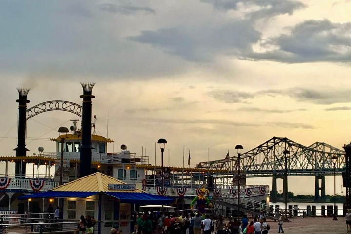 Riverboat docked near a bridge at dusk with a crowd gathered on the shore.