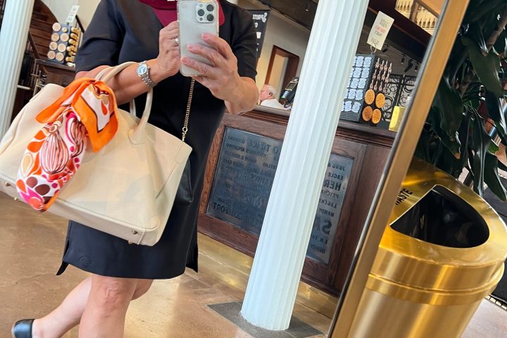 Woman taking a mirror selfie in a room, holding a red cap and a white bag with a colorful scarf.